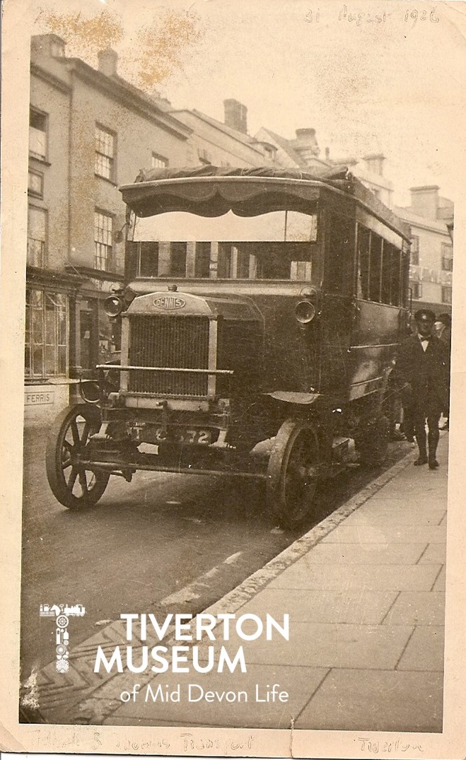 An old fashioned bus parked next to a pavement with a man in a driver's outfit and hat standing next to it. There are buildings in the background.