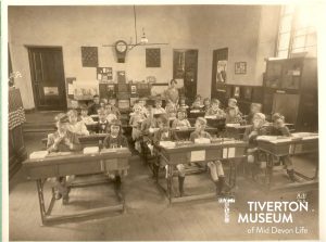 A classroom full of small children sitting at wooden desks looking at the camera. They all look very serious. The photo is in black and white.