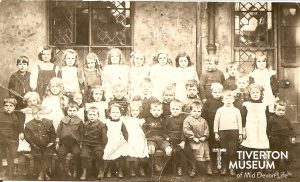 A group of children in pinafore dresses and other old fashioned clothing looking at the camera. They are all straight faced. 