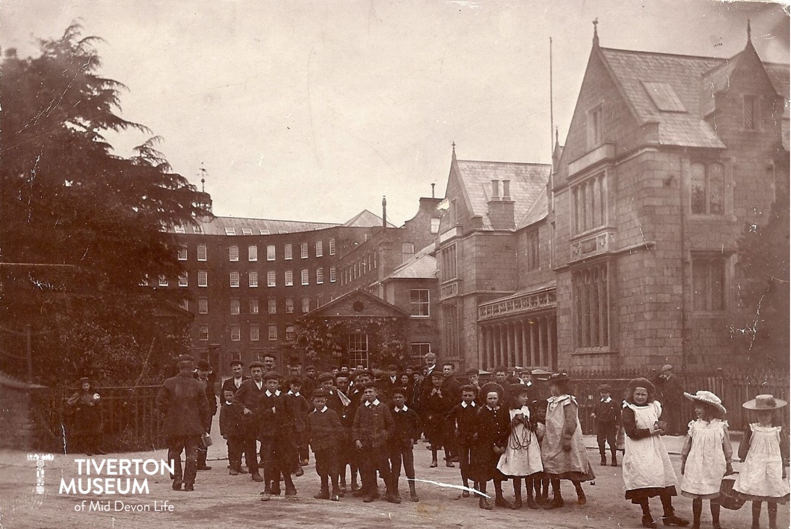 A large factory building with small children in old fashioned clothing standing in front. The photo is black and white.