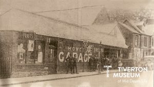 A motor garage with 'Howe's Garage' painted in white across the front of the building. There are some men gathered in front of the building, advertising signs and posters in the windows and a car in the background, but the photo is very grainy so it's difficult to pick out detail.