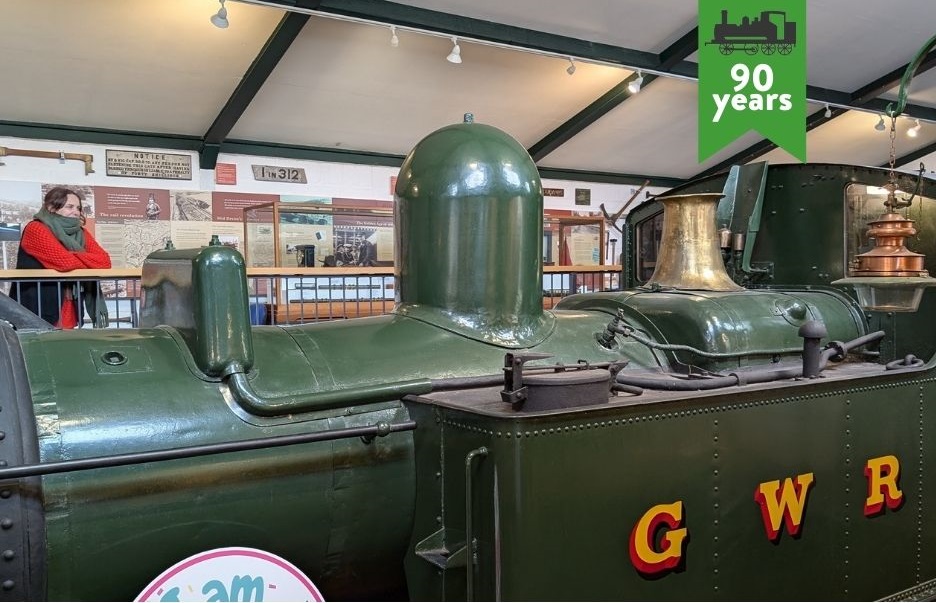 A large green steam engine with the letters GWR on the side takes up most of the shot. There is a woman leaning over the railing behind the engine looking at it in a contemplative way.