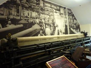 A large machine with a roller loaded with lace netting stands in a room. In front of the machine is a sign which reads 'Bobbinet lace machine'. The wall behind the machine is covered with a large black and white photo showing similar machines in their original setting at the factory with male factory workers, some of who look like they can't be more than about 12, looking at the camera.