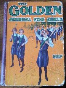 Front cover of 'The Golden Annual for Girls, 1927'. Below the title of the book there is a colour illustration of some girls in old fashioned PE kits holding hockey sticks. They look happy and smiling.