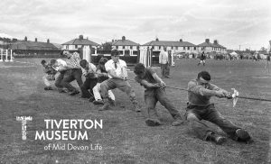 8 men are pulling on a taut rope in a playing field. The photo is in black and white and the men are wearing long trousers, shirts and jumpers, most with boots. They are clearly putting a lot of effort in!