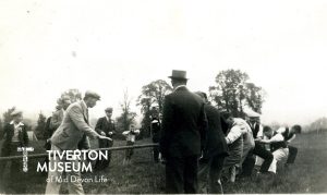 A group of men are pulling on a taut rope and there are lots of other men and boys watching on. The me are wearing suits, but with jackets removed. 