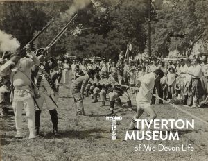 Men in sports kit (rugby style shirts and shorts) pulling on a rope. There is a large crowd gathered watching and, on the left of the group of men, there are two men in old fashioned military uniforms firing long guns into the sky.