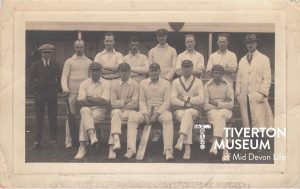 A group of men in cricket whites looking directly at the camera. The front row are seated, all wearing soft caps, with the middle man holding a cricket bag with shin pads on. The back row of 6 are standing and there is another man dressed as an umpire in a long white coat over a suit. 