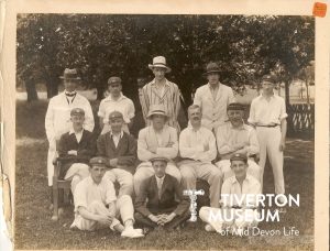A group of men posed for the photo looking directly at the camera. All in cricket whites or suits, wearing hats. They are seated or or standing, underneath some trees.
