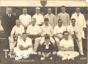 A group of men, some seated, some standing, wearing cricket whites, grouped for the photo. Some have shin pads and there is one younger boy sitting in the middle front